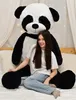 A young woman sitting on a bed next to a very large stuffed panda. Produktbild-Vorschau
