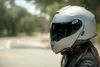 A person wearing a silver motorcycle helmet with a dark visor, outdoors on a road with trees in the background. Náhled obrázku produktu