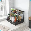 A two-tier black metal spice rack filled with various bottles and jars of spices and condiments, placed on a white marble countertop next to a window with blinds. Produktbild-Vorschau