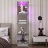A tall, narrow glass display cabinet with LED lighting, showcasing figurines and books, placed next to a bed and a small side table with a decorative bear. Produktbild-Vorschau