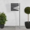 A modern mailbox with a newspaper compartment mounted on a stand, placed outdoors next to plants. Náhled obrázku produktu