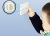 A child's hand is shown attaching a white smartphone wall mount to a blue wall. A magnified inset shows the adhesive backing of the mount. Náhled obrázku produktu