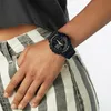 A person wearing a black G-Shock watch on their wrist, with striped pants visible. Voir les images du produit