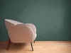 A close-up side view of a textured beige armchair with dark wooden legs against a dark green textured wall and a light wooden floor. Produktbild-Vorschau
