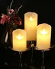 Three LED candles of varying heights are arranged on a reflective surface, with a bouquet of flowers in a vase in the background. Podgląd zdjęcia produktu