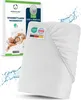 A white fitted sheet is shown diagonally, with a box of the same product in the background. Water splashes are visible around the sheet. Produktbild-Vorschau