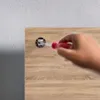 A hand holding a red screwdriver is shown inserting it into a circular fixture on a wooden surface. The background is a textured gray wall. Náhled obrázku produktu