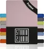 A pink fitted sheet is shown folded, with a black band across the top corner and a black panel at the bottom displaying a logo. Several other colored fitted sheets are stacked to the left and right. Produktbild-Vorschau