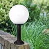 A black garden lamp with a white spherical top stands on a stone wall, with green foliage in the background. Voir les images du produit