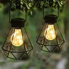 Two solar garden lanterns with diamond-shaped metal cages and warm white LED bulbs hang from tree branches. Produktbild-Vorschau