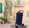 A navy blue shopping trolley with red accents stands on a cobblestone street in front of an old building with green shutters and climbing plants. Podgląd zdjęcia produktu