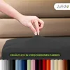 A hand is shown stretching a dark grey fitted sheet over a mattress. Below the sheet, a row of colored swatches is displayed. Produktbild-Vorschau