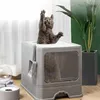 A gray cat is playing with its paws sticking out of a gray litter box, with a plant in a pot in the background. Produktbild-Vorschau