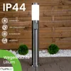 A stainless steel bollard light with a white illuminated top, set in an outdoor garden environment with wooden fencing and gravel. Produktbild-Vorschau