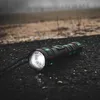 A black and green flashlight is lying on a dark, textured surface, possibly asphalt, with a blurred background of a field and distant structures. Produktbild-Vorschau