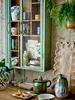 A rustic kitchen scene featuring a green display cabinet filled with ceramic mugs, vases, and artwork, with various kitchen items displayed on a wooden table in the foreground. Produktbild-Vorschau