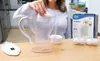 A person is inserting a water filter into a water pitcher. The product packaging and extra filters are visible on the table. Náhled obrázku produktu