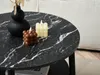 A close-up of a round coffee table with a black marble top, featuring two decorative candles and a black vase with dried flowers. Produktbild-Vorschau