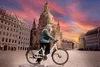 A woman is riding a white e-bike in a European city square with a large church in the background during sunset. Produktbild-Vorschau