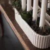 A close-up view of a white ceramic candle holder with a ribbed texture, filled with greenery and dark berries, holding white pillar candles. The holder sits on a wooden surface. Produktbild-Vorschau