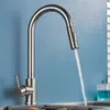 A stainless steel kitchen faucet with a pull-out spray is shown in operation, with water flowing into a sink. A blue glass with blue liquid is visible in the background. Produktbild-Vorschau