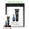 A hand holding a smartphone is taking a picture of a rotating display stand with two cosmetic bottles on it, inside a light box. Náhled obrázku produktu