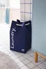 A navy blue laundry basket with the word "Laundry." printed on it, placed on a tiled floor next to a light blue cabinet in a bathroom setting. A light blue towel is visible on the edge of a bathtub in the background. Náhled obrázku produktu