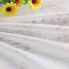 A close-up view of a white sheer fabric with a gold leaf pattern, with yellow sunflowers in the background. Produktbild-Vorschau