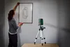 A woman is hanging a framed picture on a wall, using a laser level mounted on a tripod to ensure it is straight. The laser projects red lines on the wall. Produktbild-Vorschau