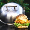 A burger is placed next to a stainless steel grill lid with a logo. Anteprima dell’immagine prodotto