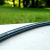 A close-up shot of a garden hose lying on a light gray surface, with a blurred green lawn and trees in the background. Voir les images du produit