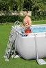 A man is standing in a rectangular swimming pool, holding onto the ladder. The pool is on a grassy lawn with trees and bushes in the background. Produktbild-Vorschau