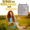 A woman is sitting in a grassy field, reading a book and holding a glass of wine. A silver insulated seat cushion is shown in the upper right corner. Produktbild-Vorschau