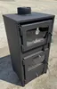 A black wood-burning stove with a glass door and a built-in oven is shown outdoors on a concrete surface. Produktbild-Vorschau
