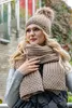 A woman wearing a beige knitted hat with a pom-pom and a matching scarf, standing outdoors with a brick wall and foliage in the background. Náhled obrázku produktu