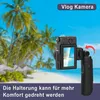 A digital camera with a flip-out screen is shown in front of a tropical beach scene with palm trees. Produktbild-Vorschau