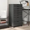 A dark gray set of drawers sits next to a desk with office supplies. Náhled obrázku produktu