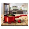 A red pot filled with food sits on a wooden table next to a tomato, with a kitchen scene in the background. Náhled obrázku produktu