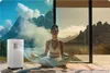A woman meditates in front of a large window with a mountain view, while a portable air conditioner is placed to her left, emitting a cool breeze. Podgląd zdjęcia produktu