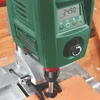 A close-up view of a green and silver drill press with a digital display and a wooden plank being drilled. Podgląd zdjęcia produktu