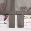 A close-up view of a grey fabric curtain with loops, showing printed floral images and text behind it. Produktbild-Vorschau