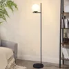 A modern floor lamp with a white glass shade is placed next to a grey armchair and a bookshelf in a living room. Anteprima dell’immagine prodotto