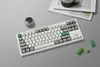 A white mechanical keyboard is placed on a light gray surface, with a few green geometric shapes and a small metal container nearby. A book with a green mug on top is visible in the background. Produktbild-Vorschau