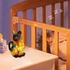 A colorful cat-shaped night light sits on a bedside table next to a crib where a baby is sleeping. Produktbild-Vorschau