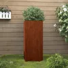 A tall, rectangular planter with a rust-like finish stands on a grassy lawn in front of a wooden fence. A green shrub is planted in the planter. A white planter with pink flowers is mounted on the fence to the left. White flowers are visible on the right. Produktbild-Vorschau