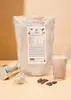 A bag of vegan chocolate protein powder is displayed next to a glass of chocolate milk, a small bowl of protein powder, and pieces of dark chocolate. A dumbbell is also visible in the foreground. Produktbild-Vorschau
