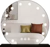 A round mirror with lights is placed on a dark surface, reflecting a sofa with pillows and a framed picture on the wall. Produktbild-Vorschau