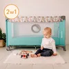 A baby is sitting on a fluffy rug next to a baby travel cot, playing with wooden toys. Produktbild-Vorschau