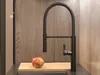A black kitchen faucet with a pull-out spray is shown in a kitchen setting, with a wooden backsplash and a cutting board with an apple in the foreground. Podgląd zdjęcia produktu