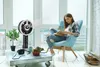 A woman is sitting in a chair reading a book next to a fan and a table with plants and a camera. Produktbild-Vorschau